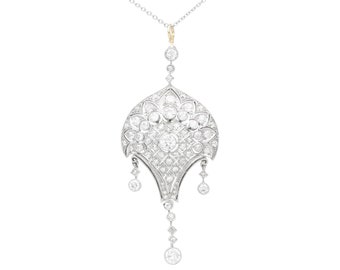 Op de afbeelding: Een zilveren en gouden ketting met een grote, sierlijke hanger met diamanten. De hanger heeft een centrale diamant die wordt omringd door kleinere diamanten in een geometrisch patroon. De ketting hangt aan een delicate ketting.