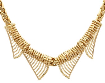 Op de afbeelding: Een gouden ketting met een delicaat, ingewikkeld ontwerp. De ketting heeft een reeks verbonden, gebogen en opengewerkte secties die een vloeiende, gelaagde look creëren.