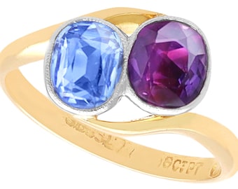 Op de afbeelding: Een gouden ring met twee ovale edelstenen, een blauwe saffier en een paarse amethist. De ring heeft een gepolijste gouden band met keurmerken. De edelstenen zijn gezet in een zilverkleurige bezel.