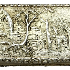 Puede incluir: Una caja rectangular de plata con un diseño ornamentado alrededor de los bordes. La tapa presenta una escena en relieve detallada de un árbol y un antiguo edificio de piedra.