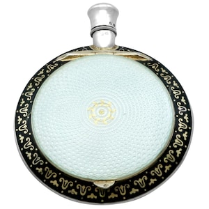 May include: A round, silver, and enamel compact with a black and gold design. The compact has a white enamel center with a gold design.