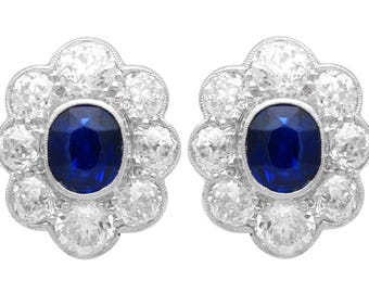 May include: A pair of elegant stud earrings. Each earring features a central oval-cut, deep blue sapphire surrounded by a halo of sparkling, round-cut diamonds. The earrings have a floral design and a silver setting.