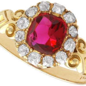 May include: A gold ring with a central oval ruby, encircled by diamonds. The ring has a decorative band with scrollwork. The ruby is a deep red, and the diamonds are set in a halo. The ring is a piece of jewellery.