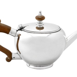 May include: A silver teapot with a wooden handle and lid knob. The teapot has a round body and a spout on the right side.