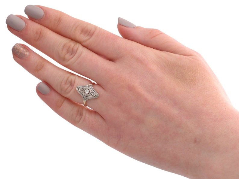 May include: A silver ring with a diamond set in an oval-shaped design. The ring is on a finger with light grey nail polish.