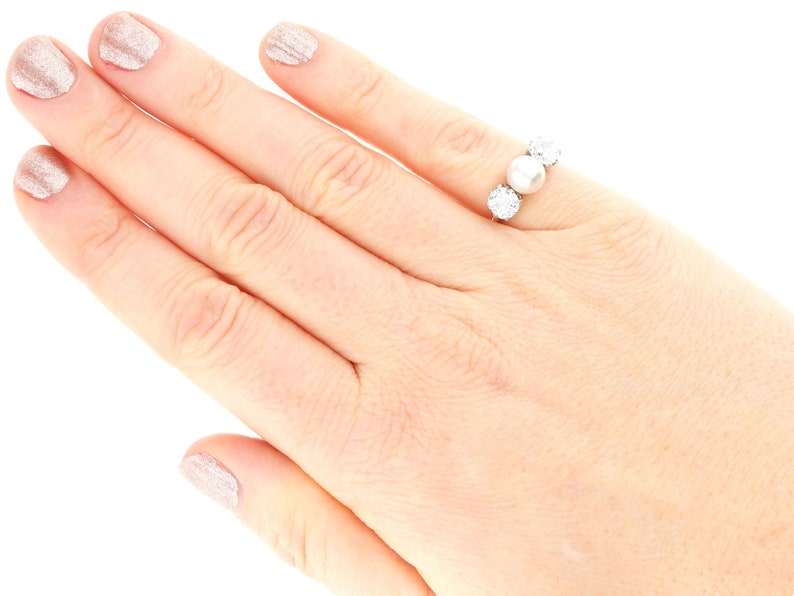 May include: A silver ring with three round white pearls set in a row. The ring is on a person's finger.
