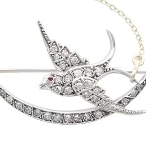 Puede incluir: Broche plateado con forma de pájaro volando, adornado con numerosos diamantes pequeños. El ojo del pájaro es una pequeña gema roja. Una barra curva y una cadena están unidas al broche.