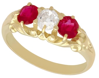 Op de afbeelding: Een gouden ring met drie edelstenen: twee levendige rode robijnen flankeren een centrale heldere diamant. De ring heeft een sierlijk, wervelend ontwerp rond de stenen en een gladde, gepolijste band.