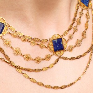 May include: A gold necklace with multiple strands featuring intricate filigree designs and square blue gemstone accents.