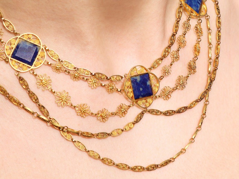 May include: A gold necklace with multiple strands featuring intricate filigree designs and square blue gemstone accents.