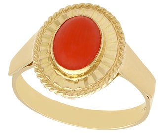 May include: A gold ring with a red coral oval gemstone set in a textured bezel. The ring has a rope-like band.