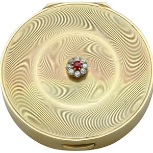 May include: A gold-toned compact with a spiral design on the lid. The center features a cluster of small diamonds surrounding a red gemstone. The compact has a hinged opening and a smooth, polished finish.