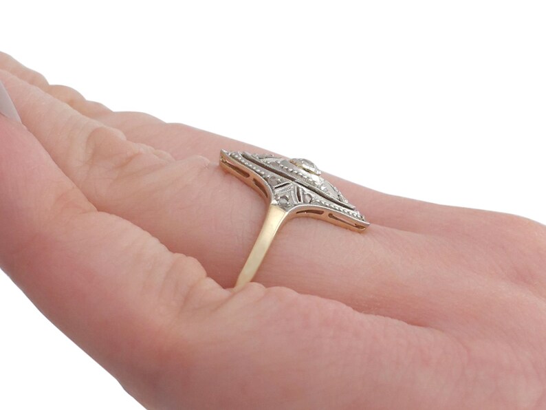 May include: A vintage diamond ring with a geometric design. The ring is set in yellow gold and features a central diamond surrounded by smaller diamonds. The ring is on a finger.