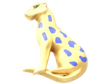 Op de afbeelding: Een broche in de vorm van een zittende luipaard, van goudkleurige metaal. De luipaard is versierd met blauwe emaille vlekken en heeft een groene edelsteen oog.