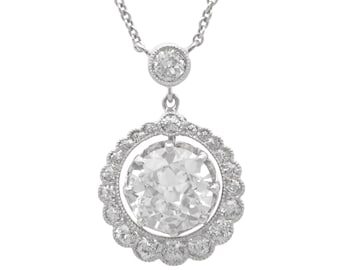 Op de afbeelding: Een zilveren hangerketting met een grote, ronde, heldere diamant omringd door kleinere diamanten. De hanger hangt aan een fijne zilveren ketting en toont een klassiek en elegant ontwerp.