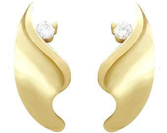 Puede incluir: Un par de pendientes de oro con un solo diamante engastado en cada uno. Los pendientes tienen forma de ala curva.
