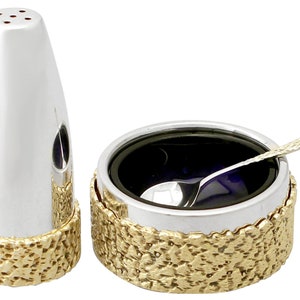 Op de afbeelding: Een zilveren zout- en peperstrooierset met een gouden, getextureerde basis. De zoutstrooier is een hoge, slanke cilinder met een klein gaatje bovenaan. De peperstrooier is een ronde kom met een klein lepeltje erin.