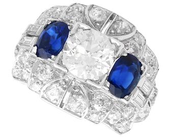 May include: A platinum ring featuring a large round diamond and two oval sapphires, all set in a geometric design with smaller diamonds surrounding the stones.