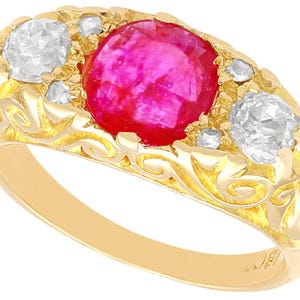 Może przedstawiać: Złoty pierścionek z dużą, okrągłą, żywą czerwoną kamieniem pośrodku, otoczonym dwoma przezroczystymi, białymi kamieniami. Pierścionek ma misternie zdobioną oprawę i obręcz z motywem kwiatowym.