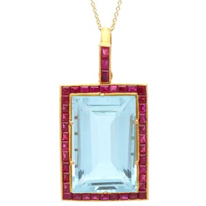 May include: A gold pendant necklace featuring a large, rectangular, light blue gemstone. The gemstone is framed by a border of small, square, red gemstones. The pendant hangs from a delicate gold chain.