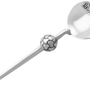 Pode incluir: Uma colher de metal prateado com um design de bola de futebol texturizado no cabo e o texto "1996" em relevo dentro da concha. O cabo é reto e afunila em direção à extremidade, enquanto a concha é redonda e possui um acabamento polido.