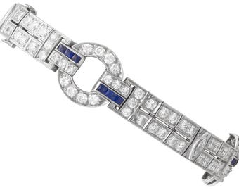 May include: A silver bracelet with a geometric design. The bracelet features clear, round gemstones and rectangular blue gemstones. The clasp is a circular design with clear gemstones. The bracelet is set against a white background.