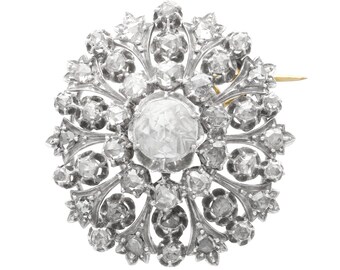 Op de afbeelding: Een zilveren en gouden broche met een groot, facetgeslepen, helder kristal in het midden, omringd door kleinere kristallen. De broche heeft de vorm van een bloem met ingewikkelde details.