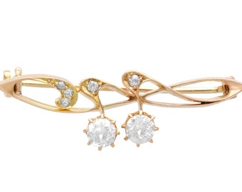 Op de afbeelding: Een gouden broche met een delicaat, vloeiend ontwerp bezet met glinsterende diamanten. De broche heeft twee ronde diamanten die aan de onderkant hangen.