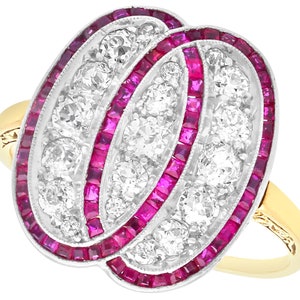 Puede incluir: Un anillo de oro con una gran piedra central de forma ovalada rodeada de diamantes y rubíes rojos más pequeños. El anillo está engastado en un diseño delicado e intrincado.