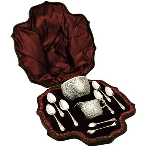 Könnte beinhalten: Ein antikes silbernes Teeservice, einschließlich Sahnekännchen, Zuckerdose und Löffel, präsentiert in einem bordeauxroten, samtgefütterten Etui. Die Silberteile weisen verzierte, gravierte Muster auf. Das Etui hat einen dekorativen, wellenförmigen Rand.