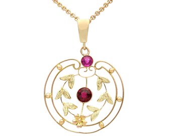 Könnte beinhalten: Ein Goldanhänger mit einem kreisförmigen Rahmen, verziert mit zarten goldenen Blattmotiven und einer kleinen goldenen Blume an der Basis. Das Schmuckstück ist mit zwei runden, dunkelrosa Edelsteinen besetzt, einer oben und einer in der Mitte.
