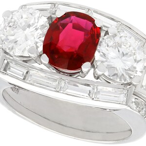 Op de afbeelding: Platina ring met een grote ovale robijn als middensteen, geflankeerd door twee ronde diamanten. De ring is verder versierd met baguette-geslepen diamanten en kleinere ronde diamanten, wat een luxueus en elegant ontwerp creëert.