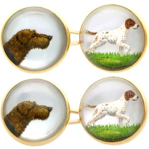 May include: A pair of gold cufflinks with glass insets. One cufflink features a painted image of a brown and white dog with black spots, standing in a field of green grass. The other cufflink features a painted image of a brown dog's head.