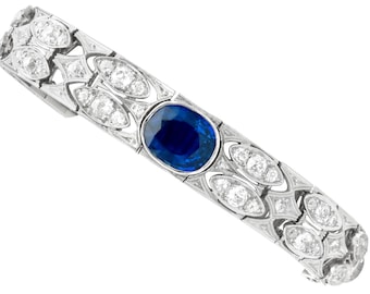 May include: A silver bracelet featuring a central oval, deep blue sapphire. The bracelet is embellished with numerous round, clear diamonds and detailed filigree work. The design showcases a repeating pattern of diamonds and decorative elements.