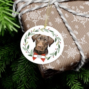 May include: A round Christmas ornament featuring a brown dog with a red bow. The dog is surrounded by a green wreath with red berries. The ornament has the name "Milo" written on a red tag.