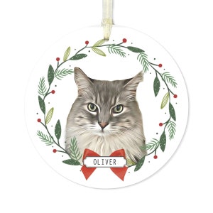 May include: A round white Christmas ornament with a green and red wreath design. A gray and white cat with green eyes is in the center of the wreath. A red bow with a white tag that says "OLIVER" is below the cat.