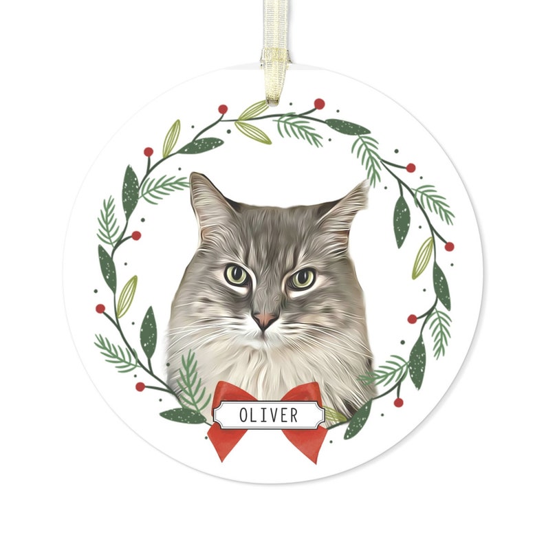 May include: A round white Christmas ornament with a green and red wreath design. A gray and white cat with green eyes is in the center of the wreath. A red bow with a white tag that says "OLIVER" is below the cat.