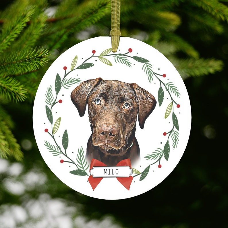 May include: A round, white Christmas ornament with a brown dog's face in the center. The dog is surrounded by a green wreath with red berries. The ornament has the name "Milo" written on a red ribbon.