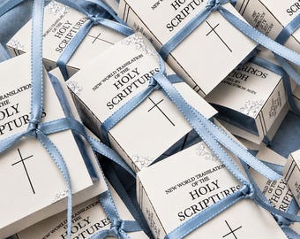 May include: A collection of small, white rectangular boxes, each tied with a light blue satin ribbon. The boxes feature the printed text New World Translation of the Holy Scriptures and a simple black cross symbol on the front cover.
