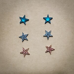 May include: Set of six star-shaped earrings. Two are blue with a glitter finish, two are silver with a glitter finish, and two are pink with a glitter finish.