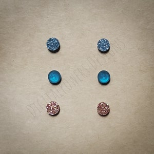 Op de afbeelding: Set van zes kleine ronde glitter oorbellen. Twee zijn met blauwe glitter, twee zijn met teal blauwe glitter en twee zijn met roze glitter.