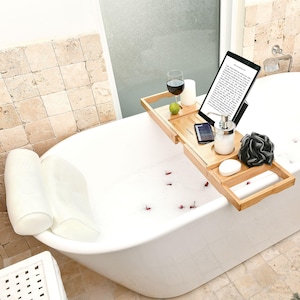 Bath gifts for women, the Monsuri bath pillow and bamboo bathtub tray caddy: the white mesh pillow at the head of an empty white freestanding tub and the tray across it, holding a tablet on the stand, a glass of red wine in its slot, a candle, a rolled towel, a phone, a soap dispenser, a bath sponge and a bar of soap.