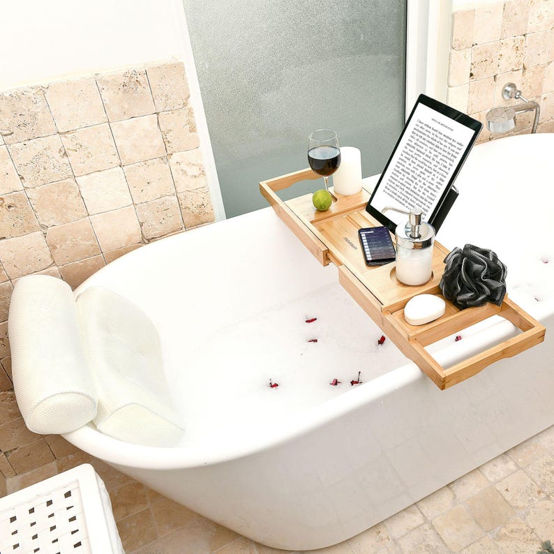 Bath gifts for women, the Monsuri bath pillow and bamboo bathtub tray caddy: the white mesh pillow at the head of an empty white freestanding tub and the tray across it, holding a tablet on the stand, a glass of red wine in its slot, a candle, a rolled towel, a phone, a soap dispenser, a bath sponge and a bar of soap.