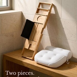 Bath pillow and bathtub tray set out of the tub: the bamboo tray leaning against a stone wall on a wooden bench and the mesh pillow beside it with its hanging hook, captioned Two pieces. One long soak.