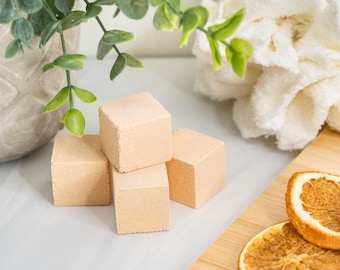 Peut inclure: Quatre bombes de bain carrées de couleur pêche clair disposées en groupe. Les bombes de bain ont une surface mate et texturée. À proximité se trouvent deux tranches d'orange séchées sur une planche en bois, une fleur de douche blanche et une plante verte dans un pot gris.