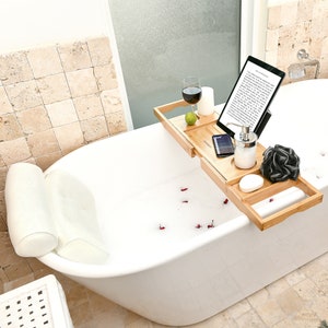 May include: A bamboo bathtub tray holds a tablet, a glass of red wine, a candle, a lime, a phone, a soap dispenser, a loofah, a bar of soap, and a rolled white towel. The tray is positioned over a white bathtub filled with water and rose petals.