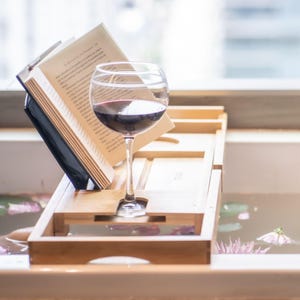 Bath pillow and bathtub tray set, the tray close up: an open book on the stand and a glass of red wine in its slot.