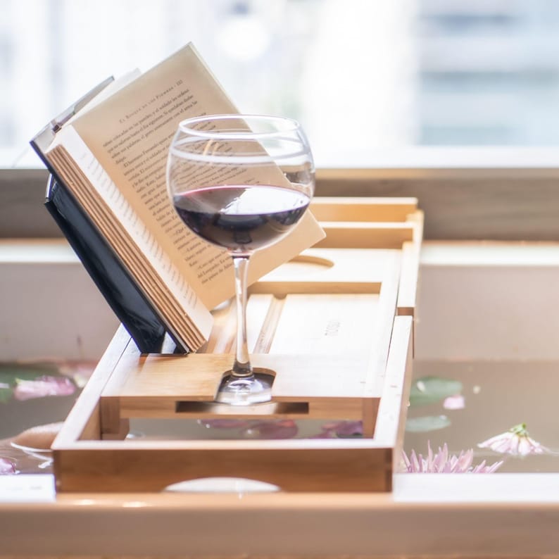 Bath pillow and bathtub tray set, the tray close up: an open book on the stand and a glass of red wine in its slot.