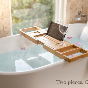 Bath pillow and bathtub tray set on a white freestanding tub: the mesh pillow at the head and the bamboo tray across the water with a tablet and a wine glass, captioned Two pieces. One long soak.