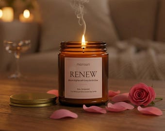 Peut inclure: Une bougie allumée dans un pot en verre ambré avec une étiquette blanche indiquant monsuri RENEW Bloom Brighter with Every Gentle Glow 198g. Un couvercle en métal doré est posé à côté, avec une rose rose et des pétales éparpillés.